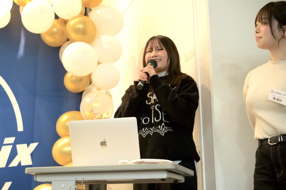 Young woman speaks into a handheld microphone at a podium with a laptop, balloons in the background.