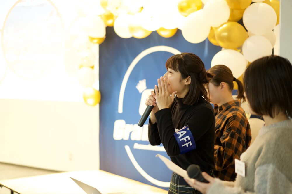 Woman wearing a blue STAFF armband speaks into a microphone at a celebration, with balloons and a blue banner behind her.