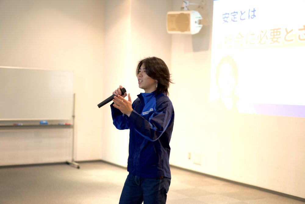 Presenter wearing a blue jacket speaks into a handheld microphone in a classroom or conference room, with a projection screen on the right.
