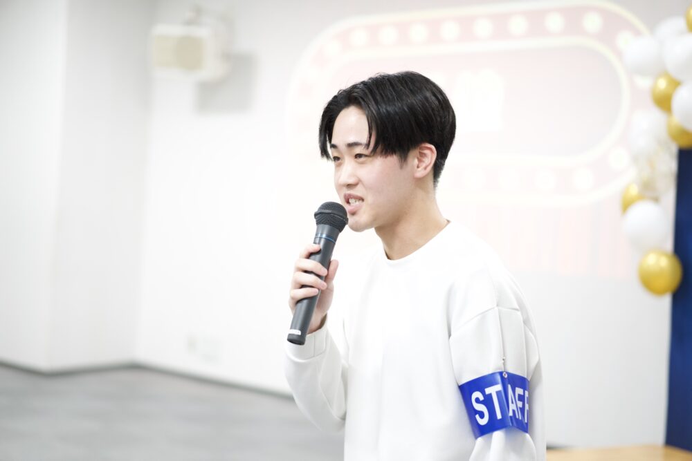 Young man in a white sweatshirt speaks into a handheld microphone at an indoor event, wearing a blue STAFF armband with balloons in the background.