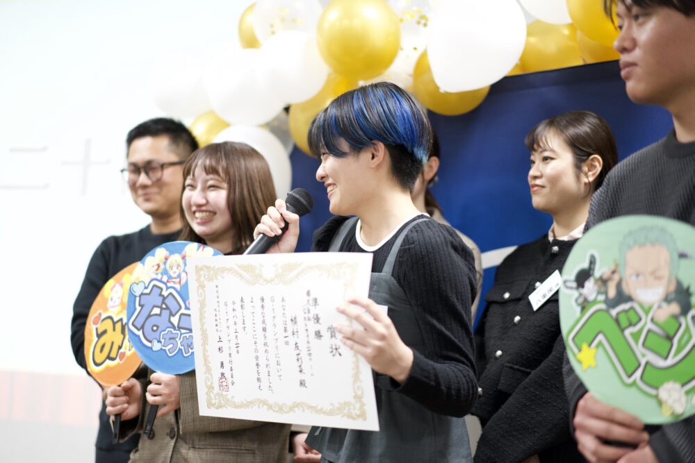 Person with blue-dyed hair speaks into a microphone and holds a certificate during a celebratory event, with balloons and colleagues in the background.