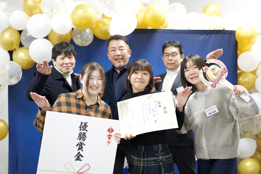 Six people posing for a celebratory photo with certificates and a large award card, framed by gold and white balloons in the background.