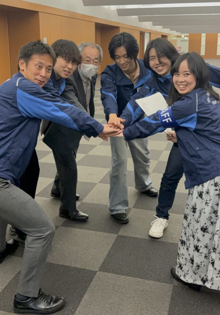Six people in blue jackets form a circle and place their hands together in the center, smiling in a hallway—team huddle or group cheer.