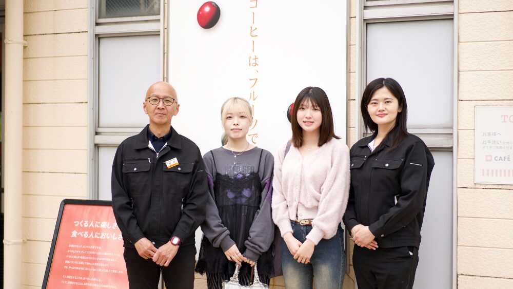 Four people standing side by side outside a building entrance, two staff in black uniforms on the ends and two casually dressed women in the middle, with a white sign featuring orange Japanese text behind them.
