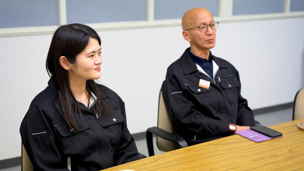 Two coworkers in black uniforms sit at a wooden conference table during a meeting in a bright room with windows in the background; they look attentive to someone off-camera.
